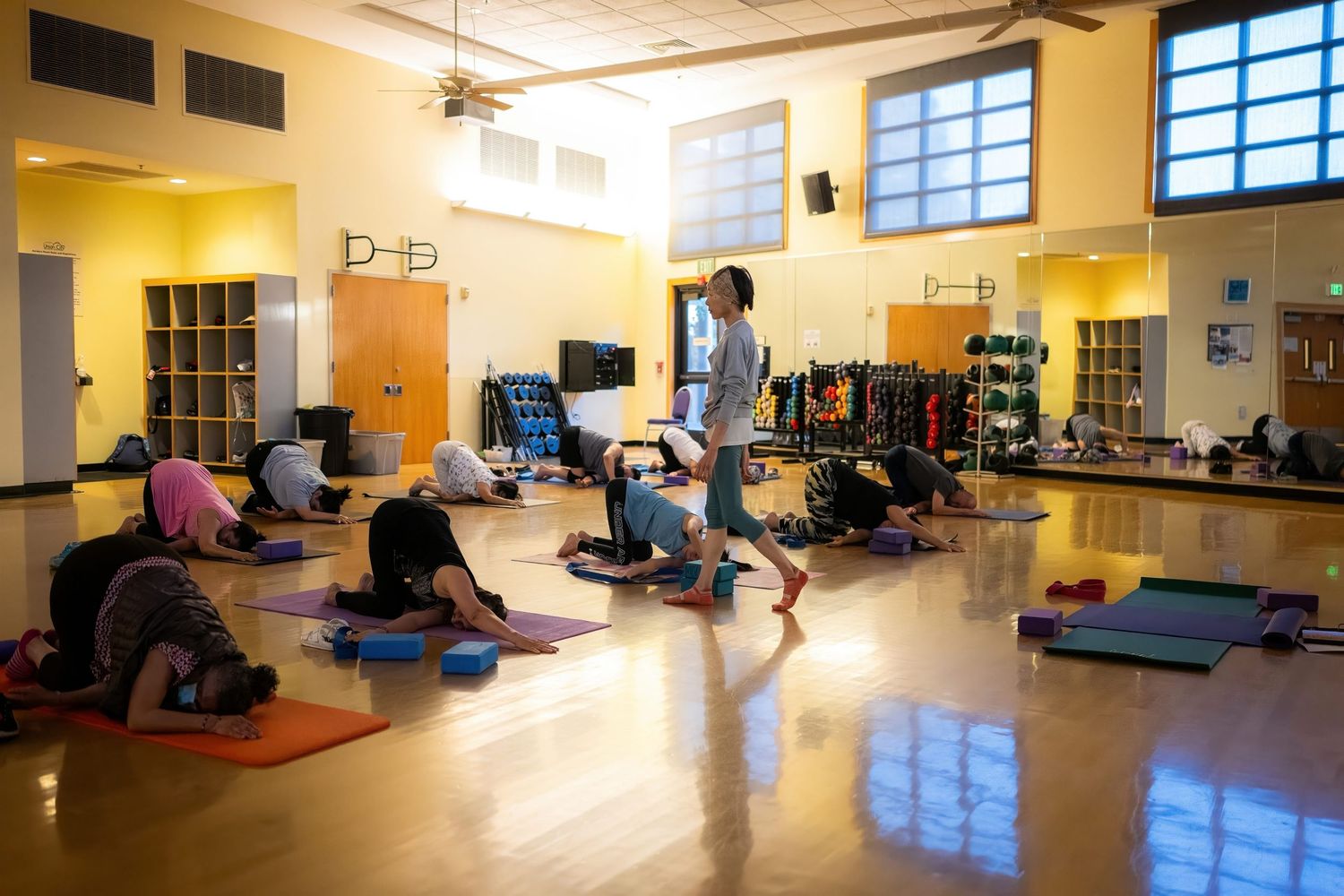 Large fitness studio with yellow walls and blue windows featuring exercise mats and equipment.