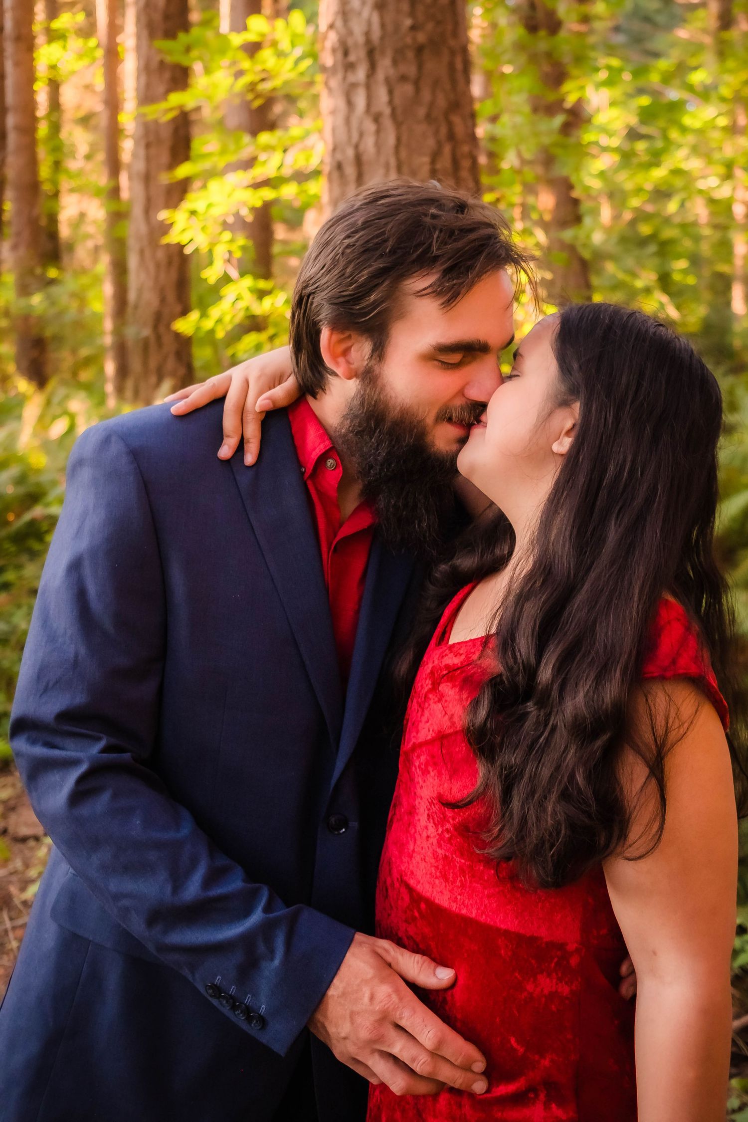 A romantic couple in navy and red formal attire share tender kisses in a sunlit forest setting.