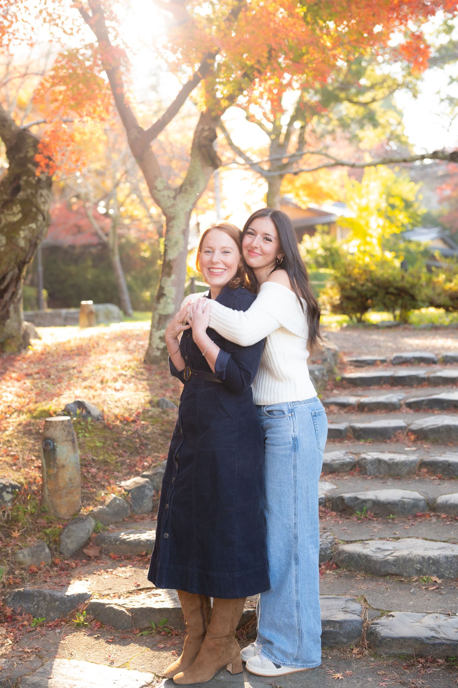 Two women embrace and smile in a sunlit autumn park with vibrant red and orange foliage.