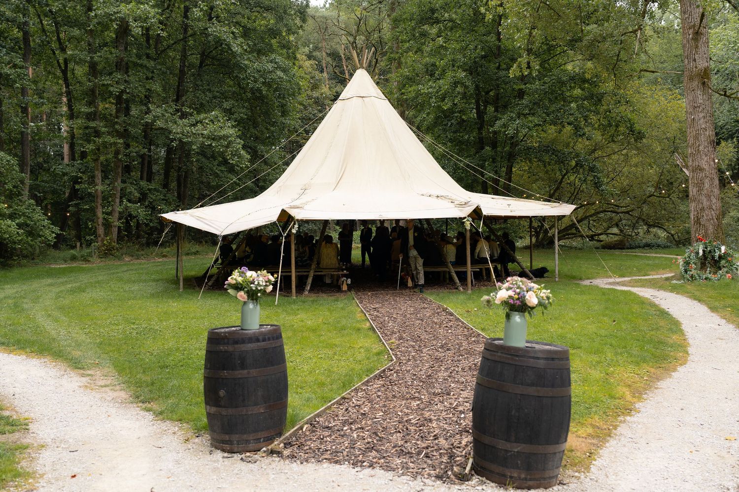 A rustic outdoor wedding venue features wine barrels and a white tent canopy along a winding gravel path.