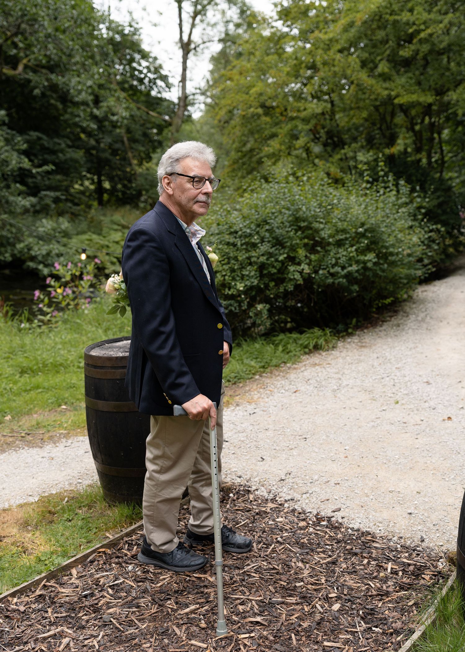 A person wearing a navy blazer and khakis stands on a gravel path in a lush garden setting, using a walking cane.