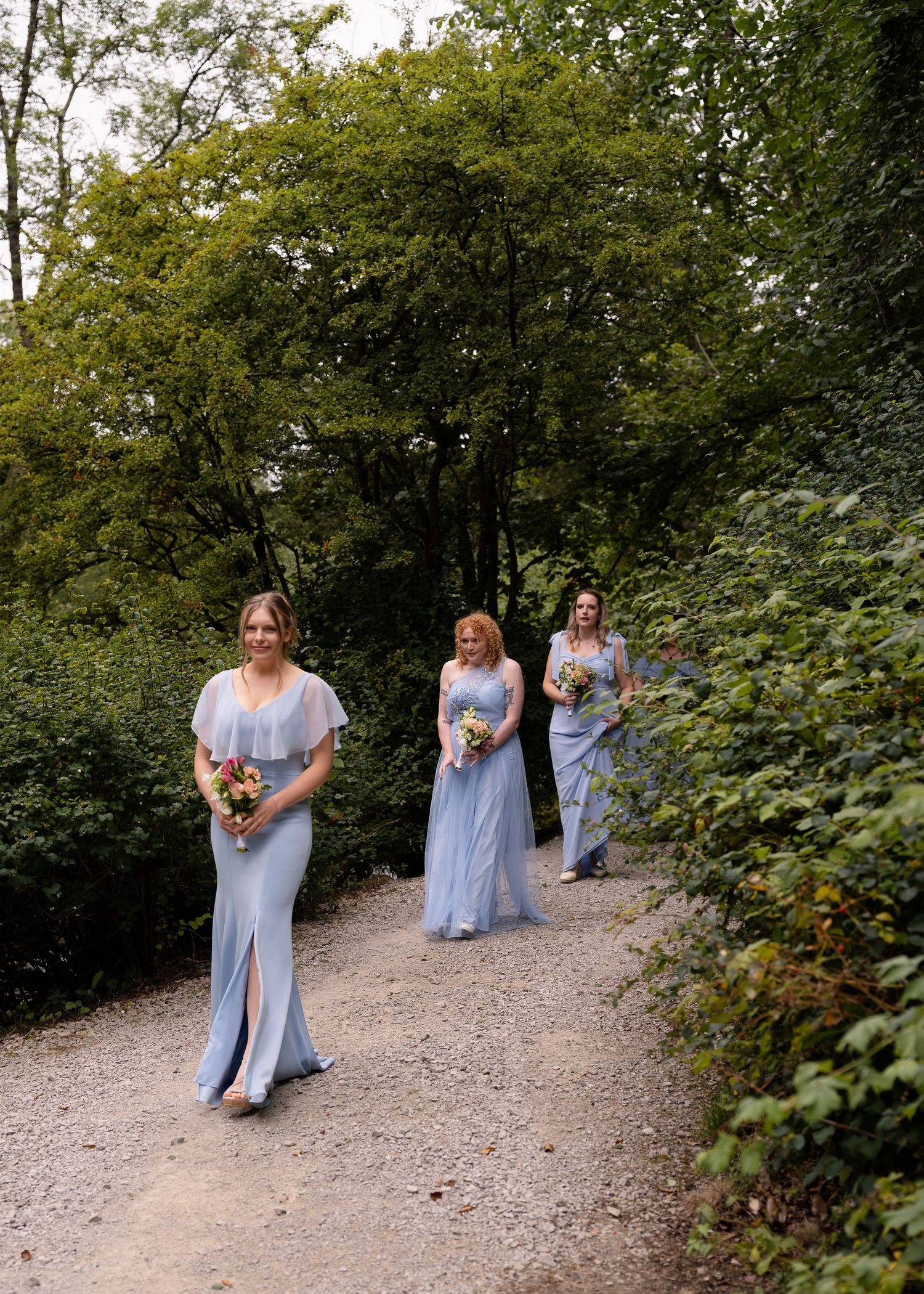 Bridesmaids in light blue dresses walk along a wooded path during a wedding procession.
