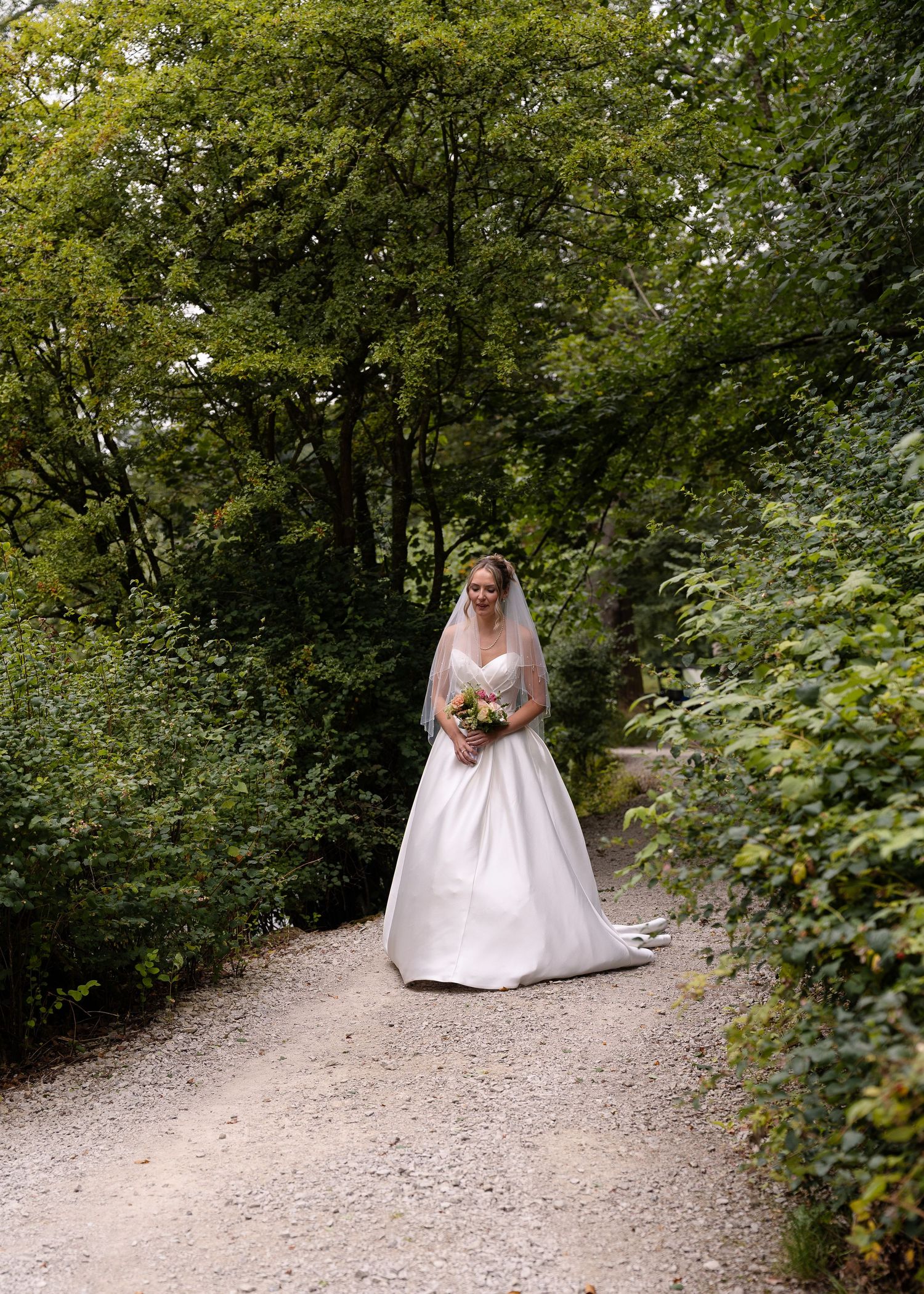 A bride in a white wedding gown stands on a winding gravel path surrounded by lush green foliage in a forest setting.