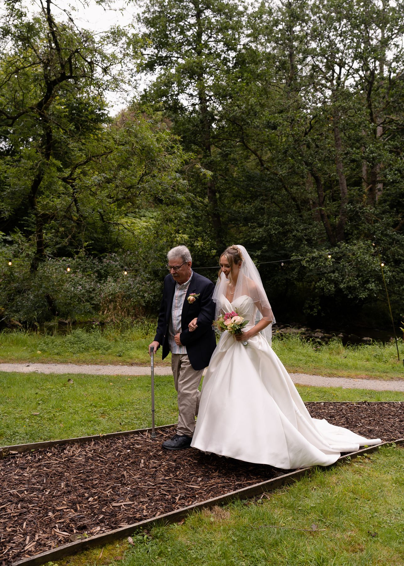 A couple in wedding attire walk along a wooden path through a lush garden setting on their special day.
