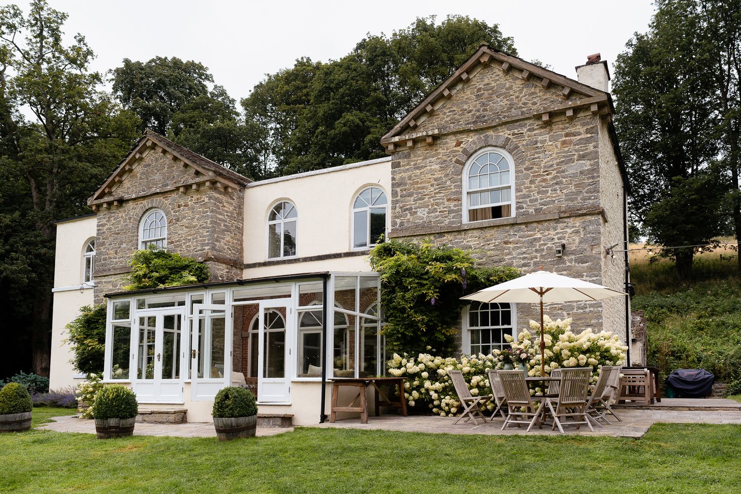 Stone cottage with glass conservatory extension surrounded by green gardens and trees in countryside setting.
