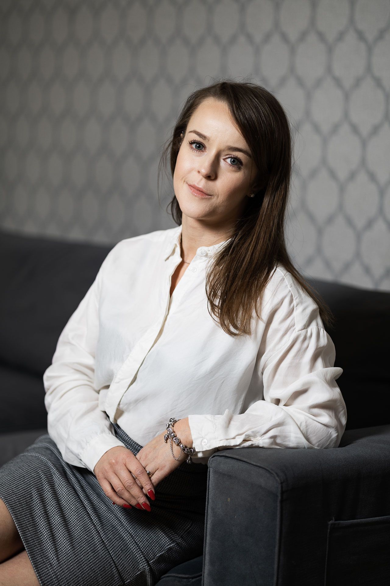 Professional portrait series showing a woman in white blouse and gray skirt reclining on dark couch against patterned wall.