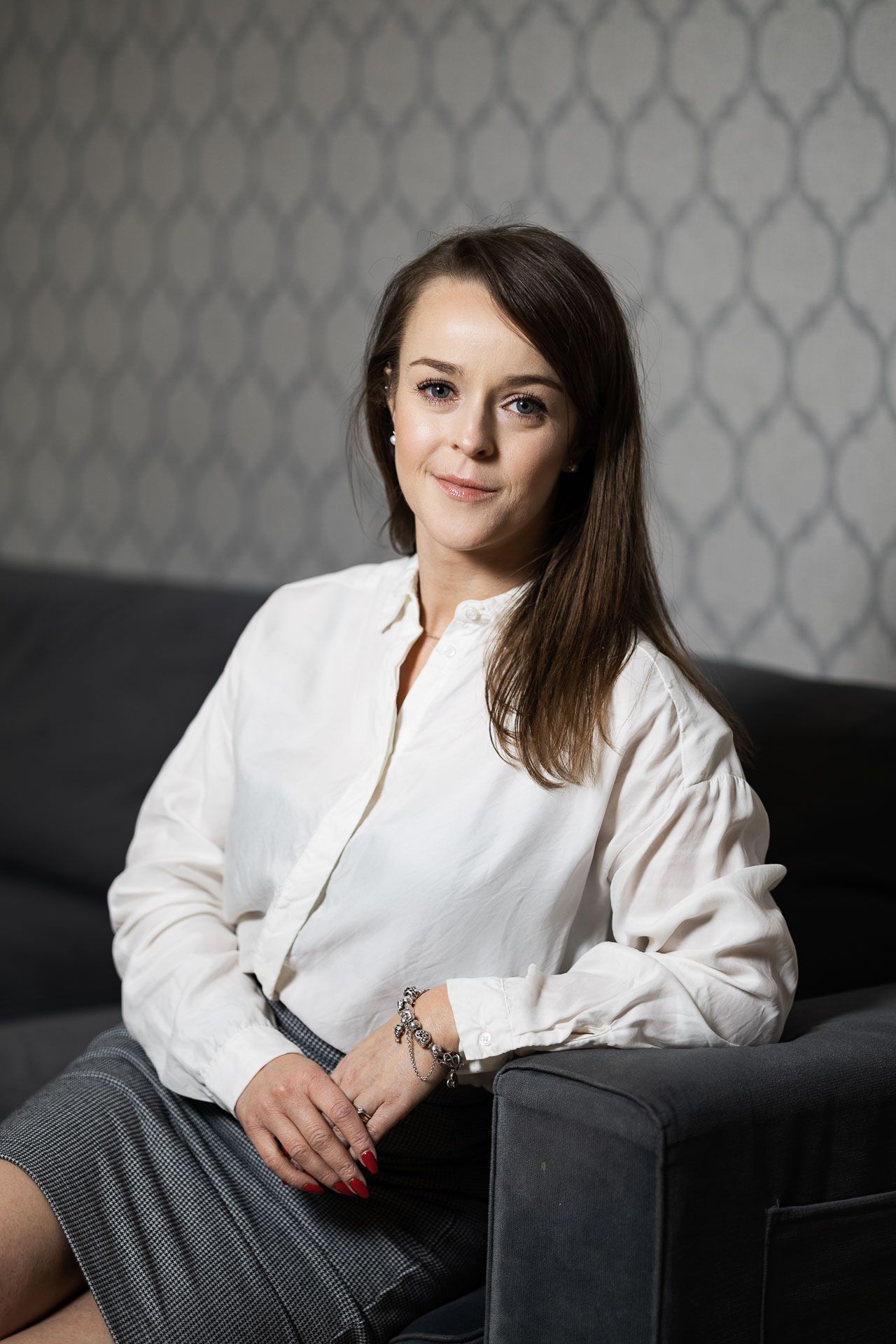 Professional portrait of someone wearing a white blouse while reclining on a dark couch against a patterned wall.
