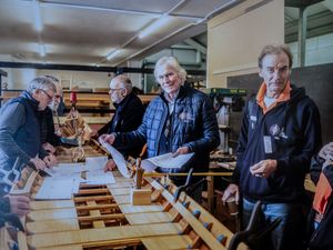 A group of people work together on a long wooden boat construction project in a workshop setting.