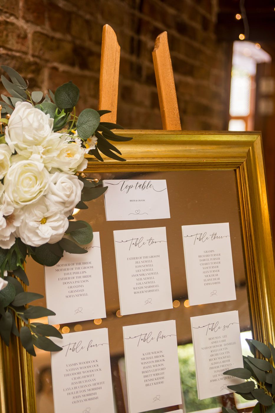 Wedding seating assignments displayed in gold frame with white floral accents.