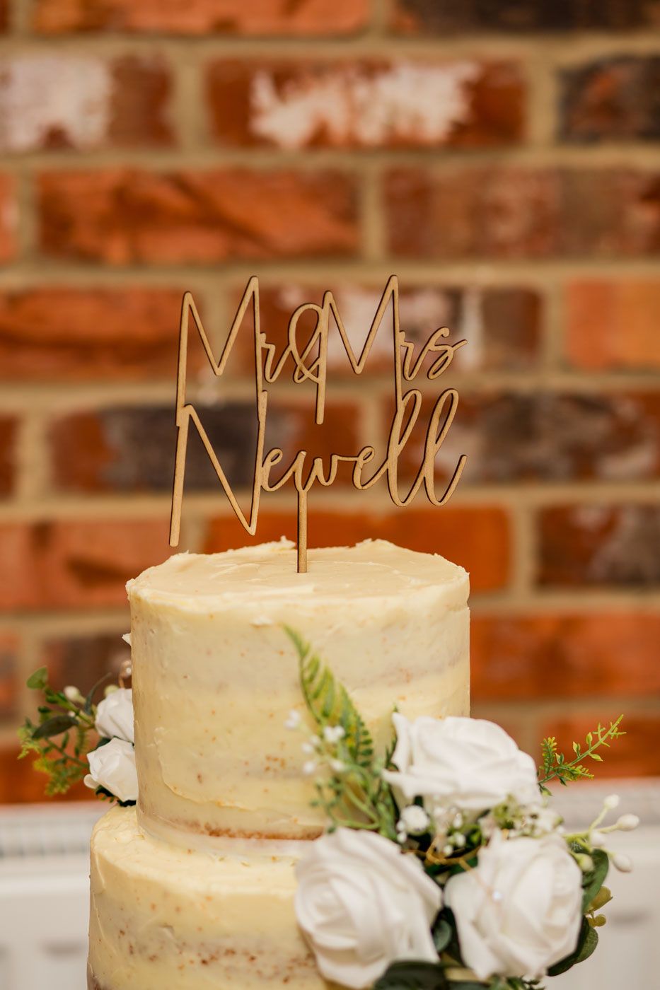 Wedding cake decorated with white roses and gold wire 'Mr & Mrs' topper against rustic brick wall.