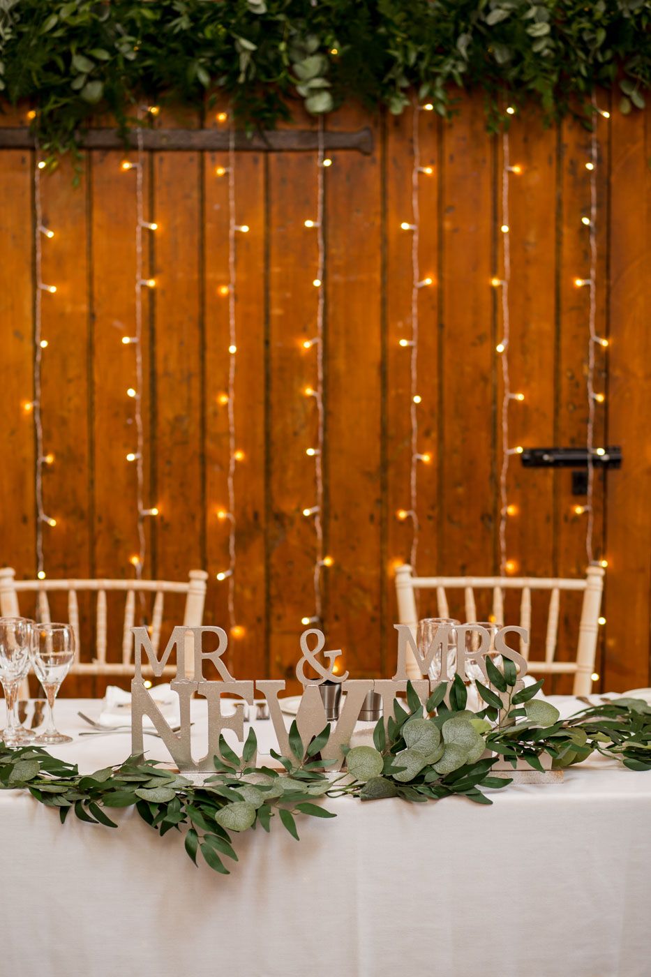 Wedding reception decor with string lights and 'Mr & Mrs' signage on wooden fence backdrop.