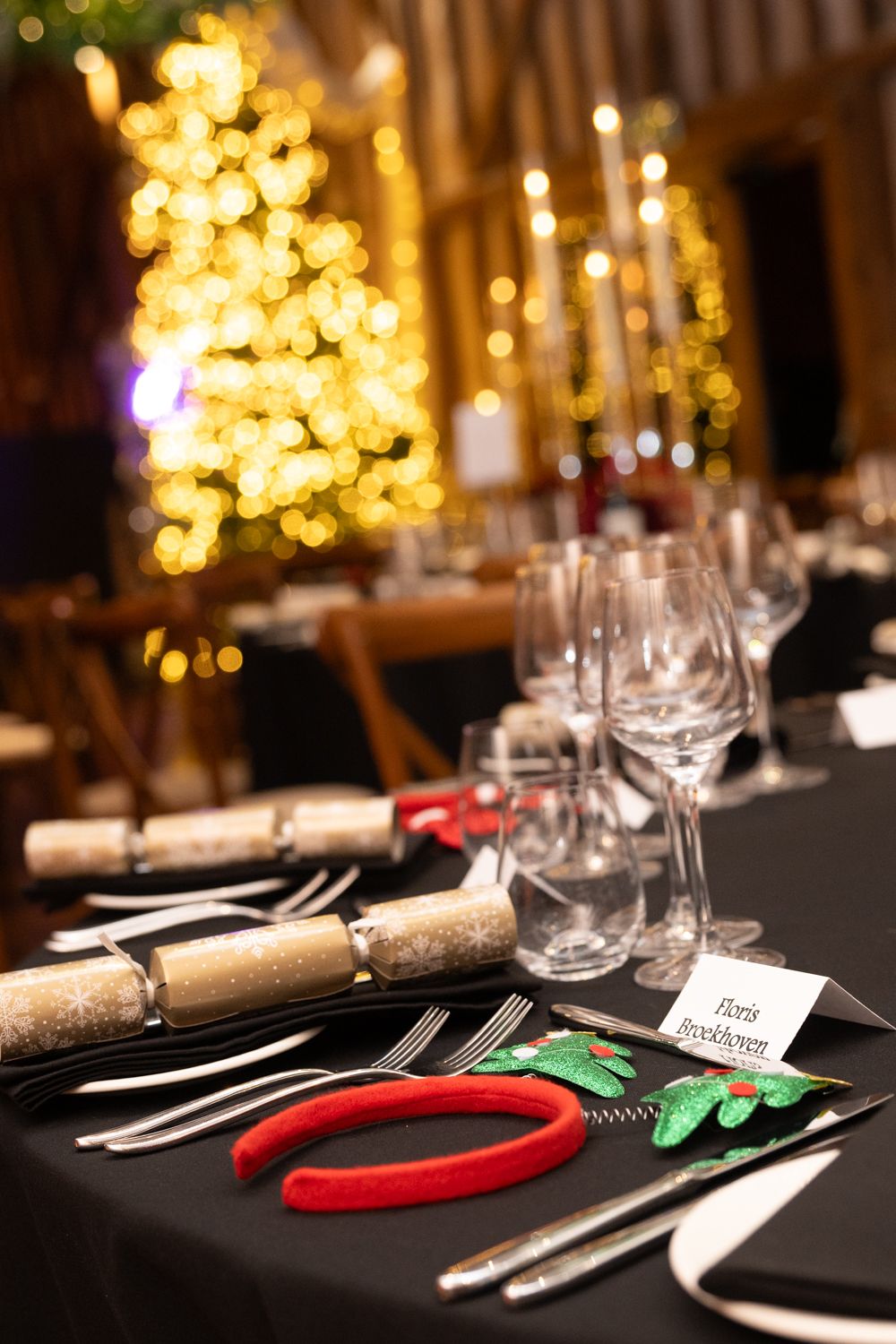 Elegant Christmas table settings with black linens, silverware, and festive crackers against blurred golden lights.