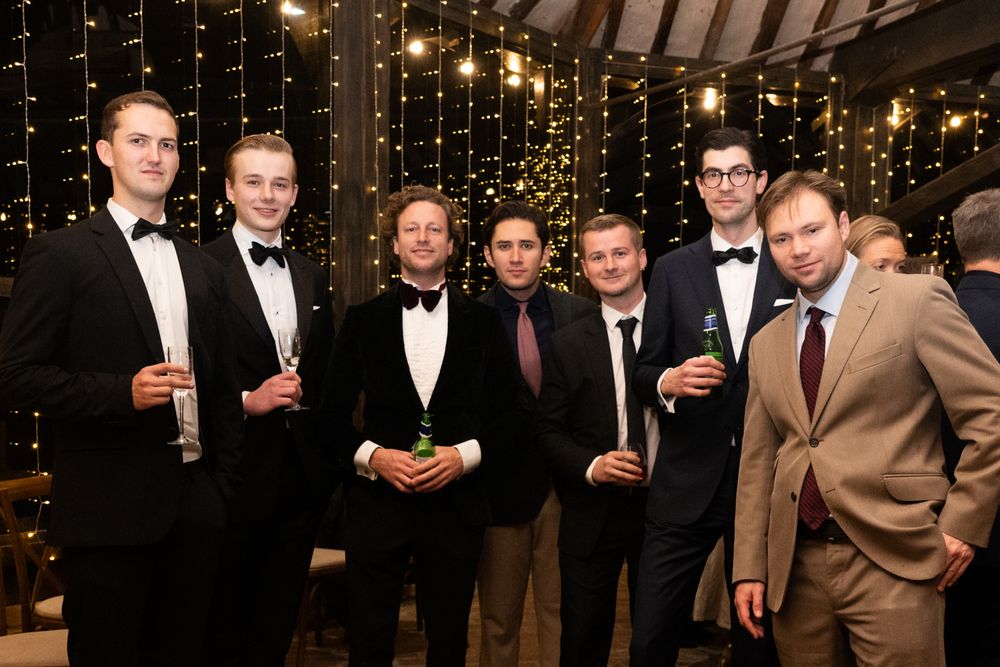 A group of well-dressed guests in tuxedos and suits gather at an elegant evening event with twinkling lights in the background.