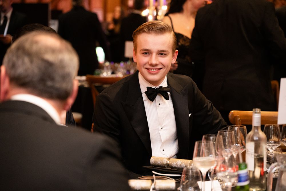 A guest in a black bow tie and tuxedo sits at an elegant dinner table with wine glasses.