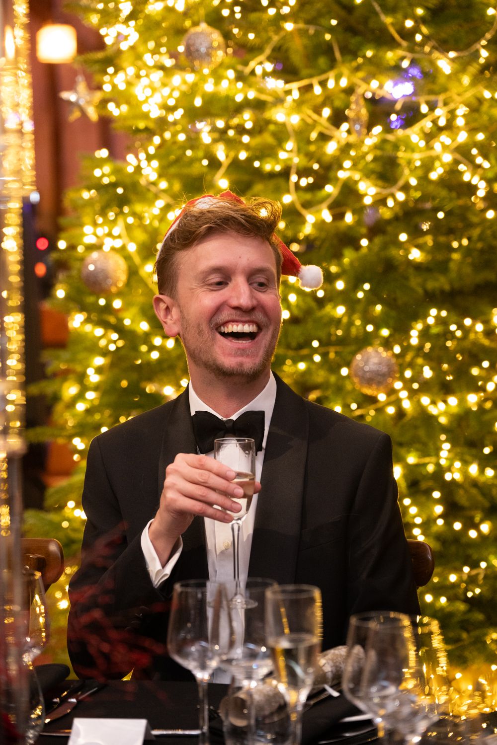 A person in a tuxedo laughs while holding a champagne glass at an elegant holiday party with a lit Christmas tree behind.