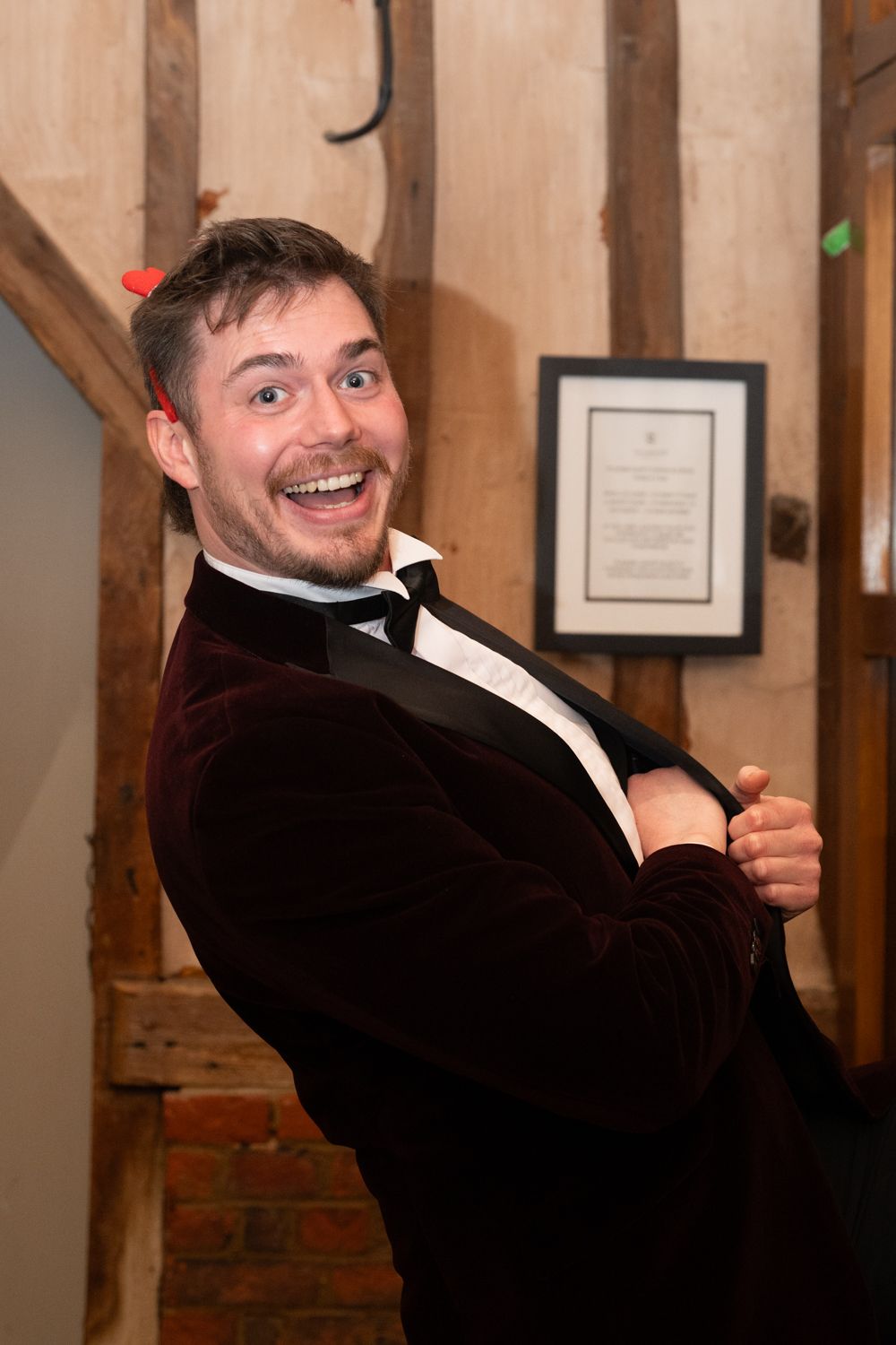 A person in a dark suit jacket smiles and gestures against a wooden wall with framed documents.