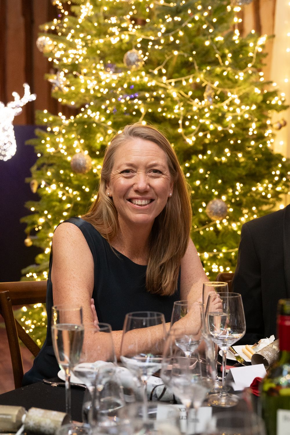 A person in a black sleeveless top smiles warmly at a formal dinner table with a Christmas tree glowing in the background.