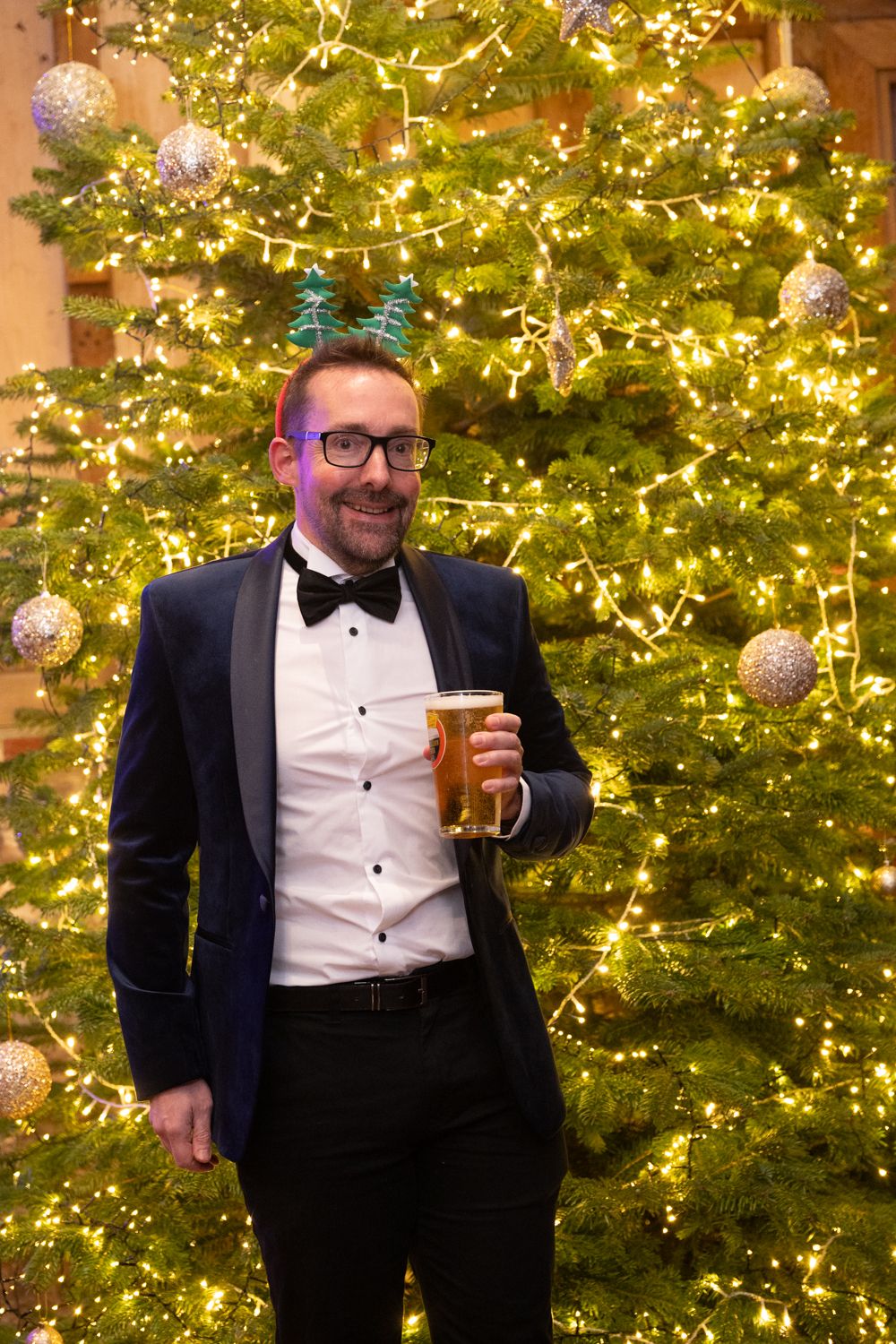 Person in formal black tuxedo and bow tie holding drink while posing in front of illuminated Christmas tree.