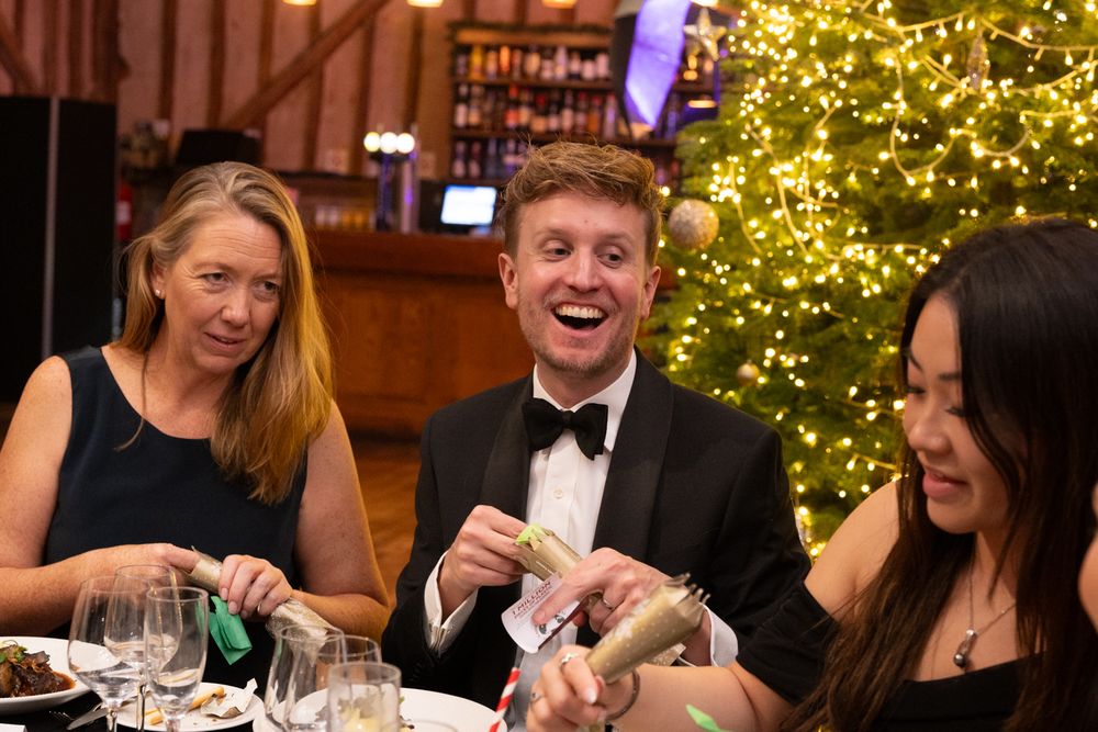 Dinner guests laugh together at a holiday party with a glowing Christmas tree in the background.