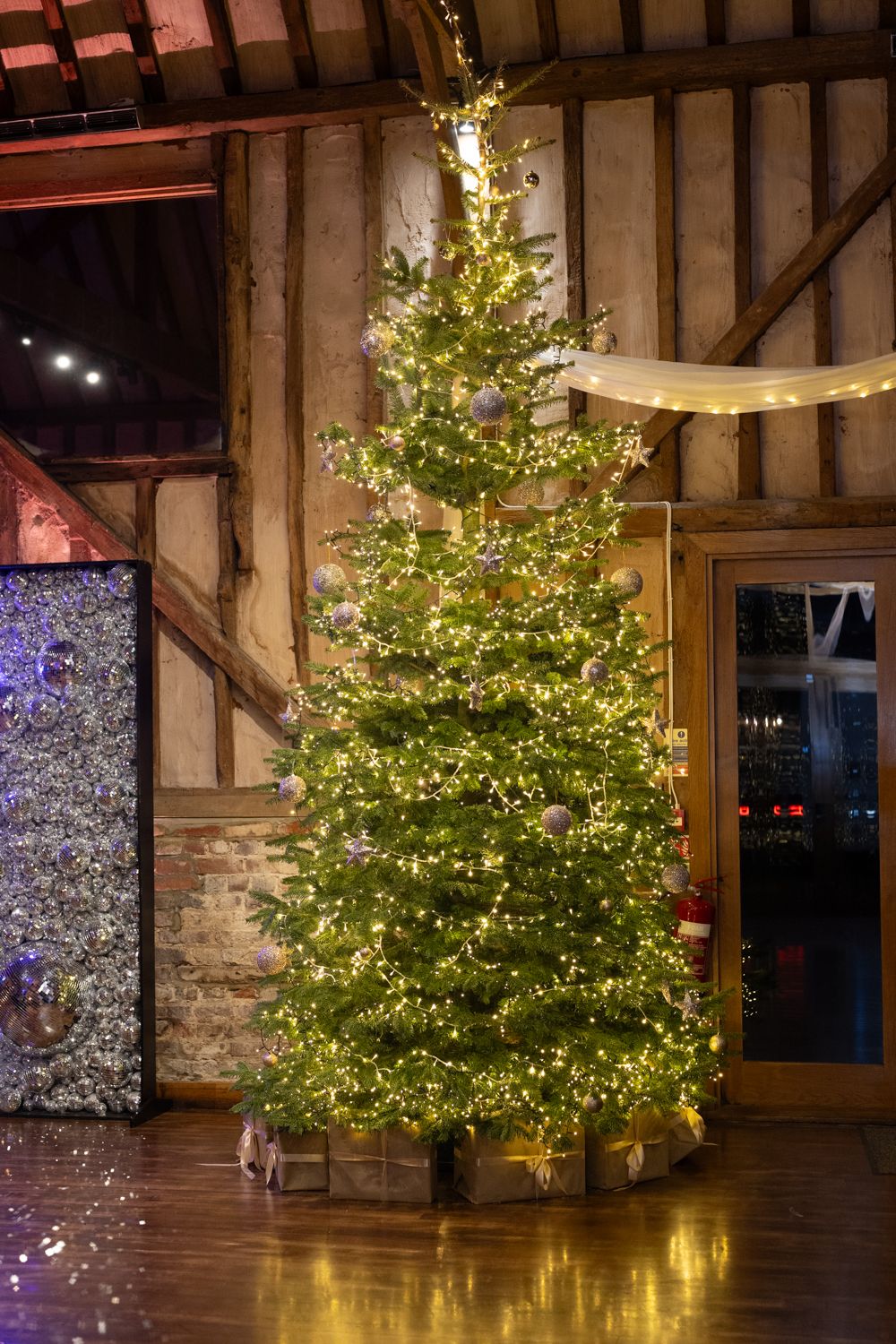 A rustic barn interior decorated with a glowing Christmas tree adorned with warm white lights against stone walls.