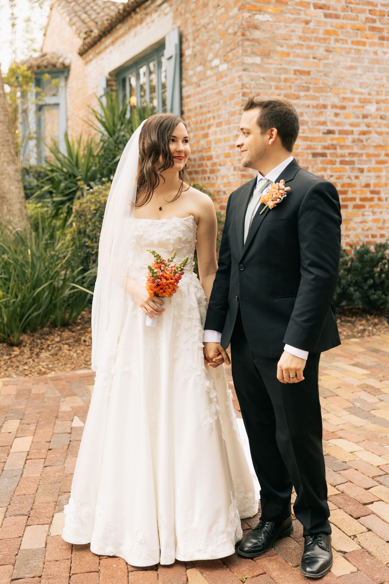 Bride in a white gown and veil holds an orange bouquet and the groom's hand on a brick path beside a historic brick building.