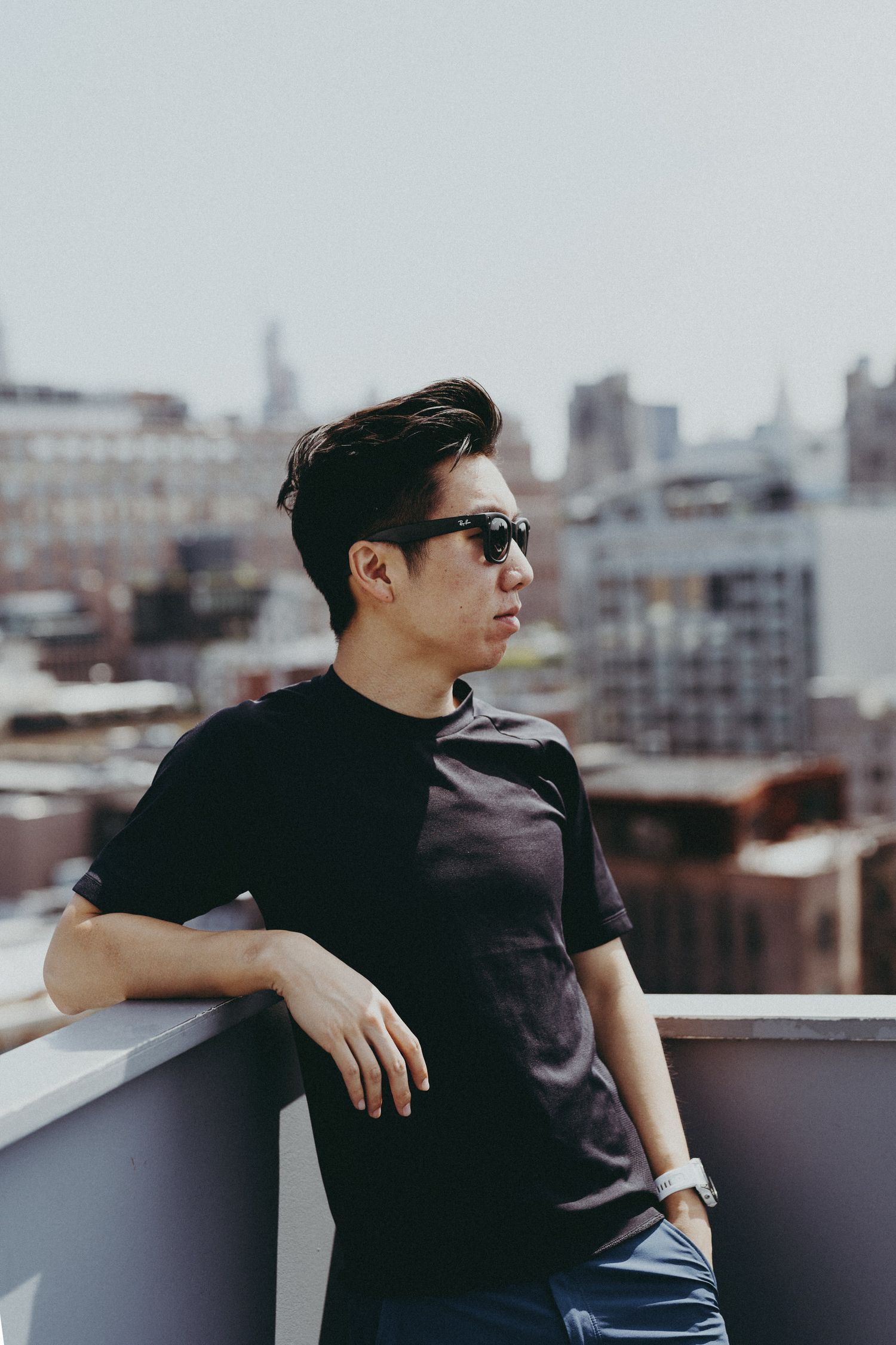 Stylish figure in black leaning on urban rooftop railing with cityscape behind.