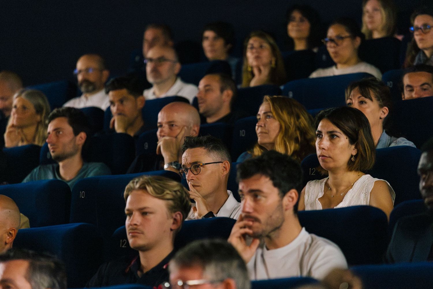 Audience watching intently in dark theater with focused expressions.