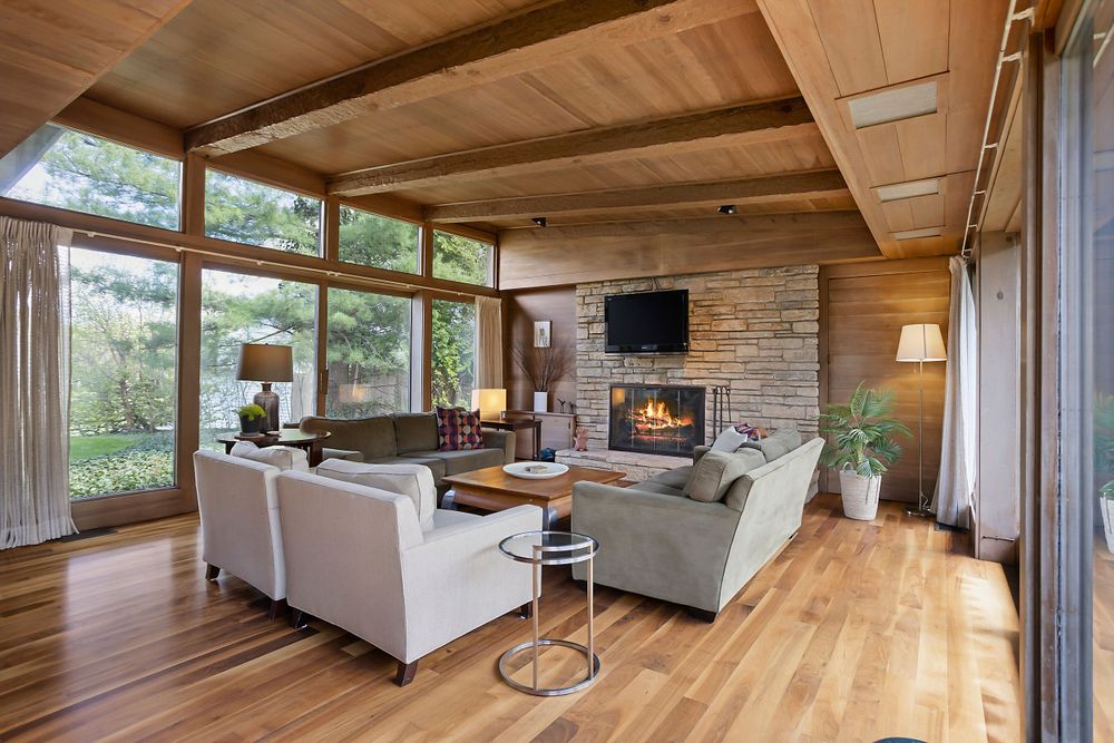 Warm living room with wood ceilings, large windows, stone fireplace, cozy seating, and lush outdoor views.