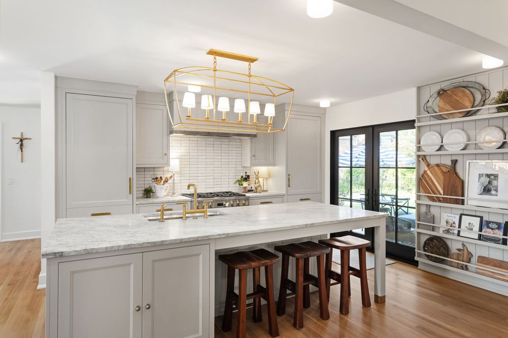 Bright kitchen with marble island, gold fixtures, elegant lighting, open shelving, and large glass doors.