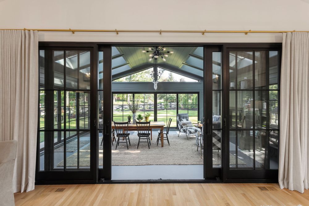 View through black-framed glass doors into a stylish sunroom with vaulted ceilings, skylights, and a dining area overlooking a green yard.