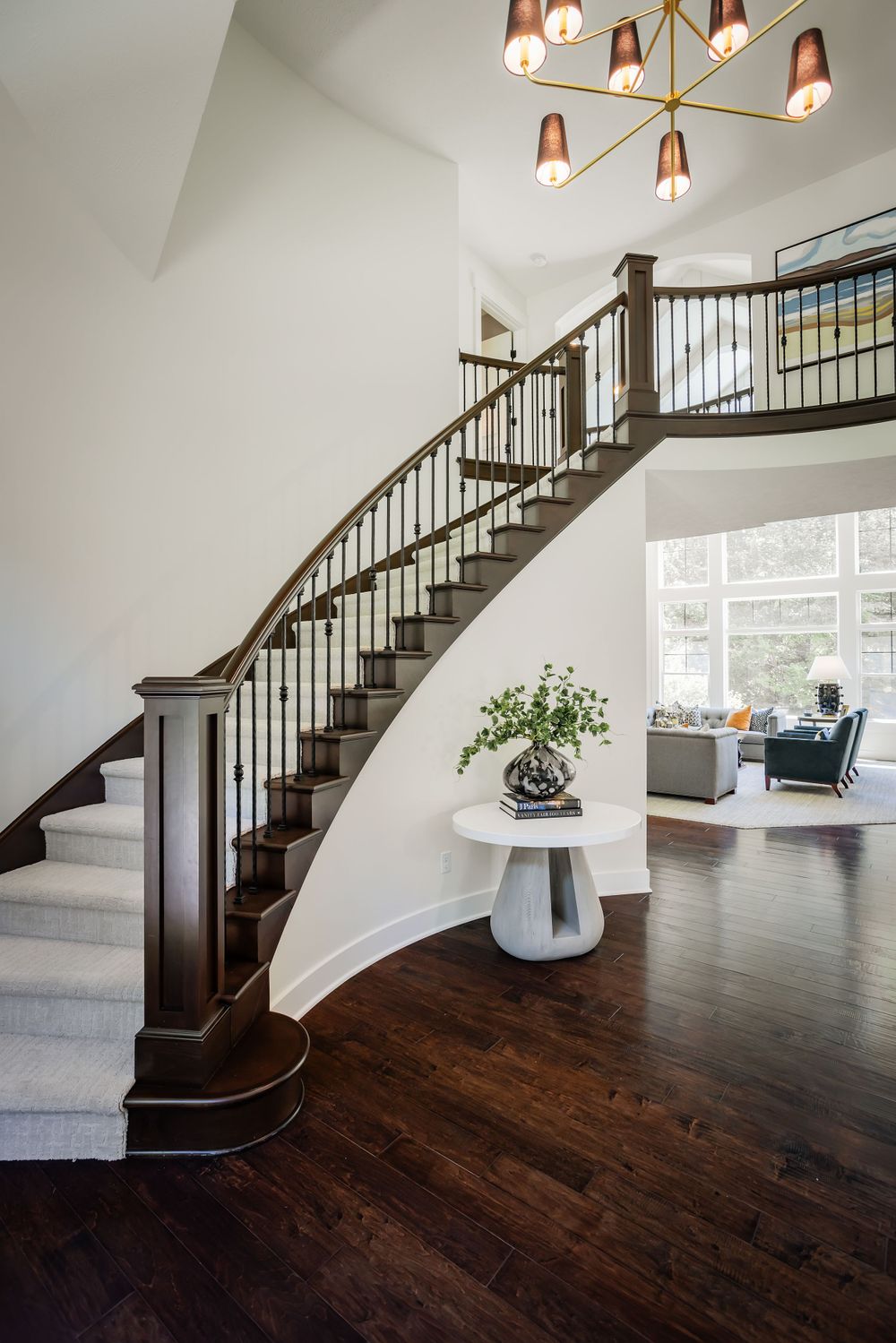 Elegant curved staircase with dark wood railing , leading to an open second-floor landing overlooking a bright living room.