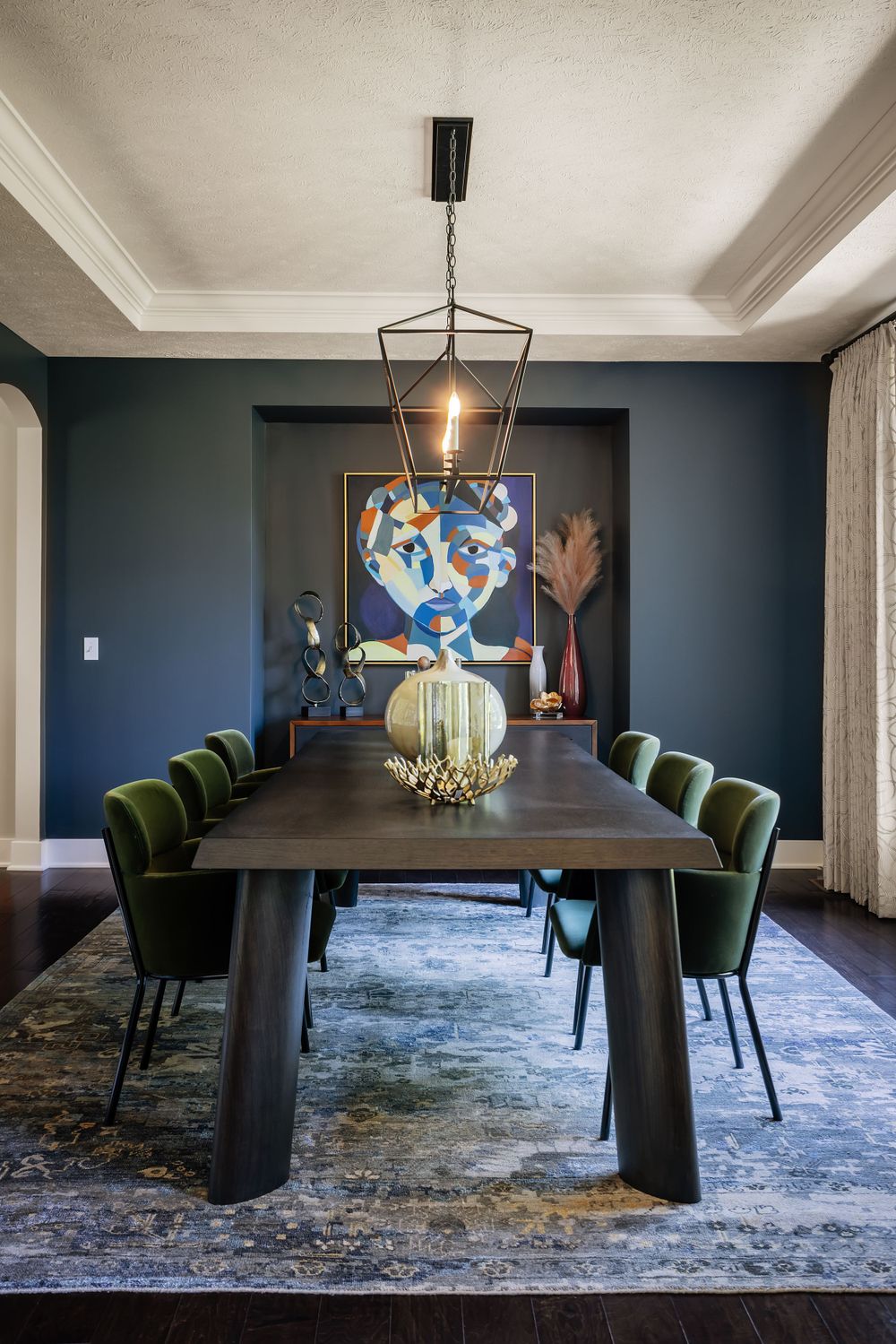 Modern dining room with a dark wood table, green velvet chairs, abstract artwork, and a geometric pendant light.
