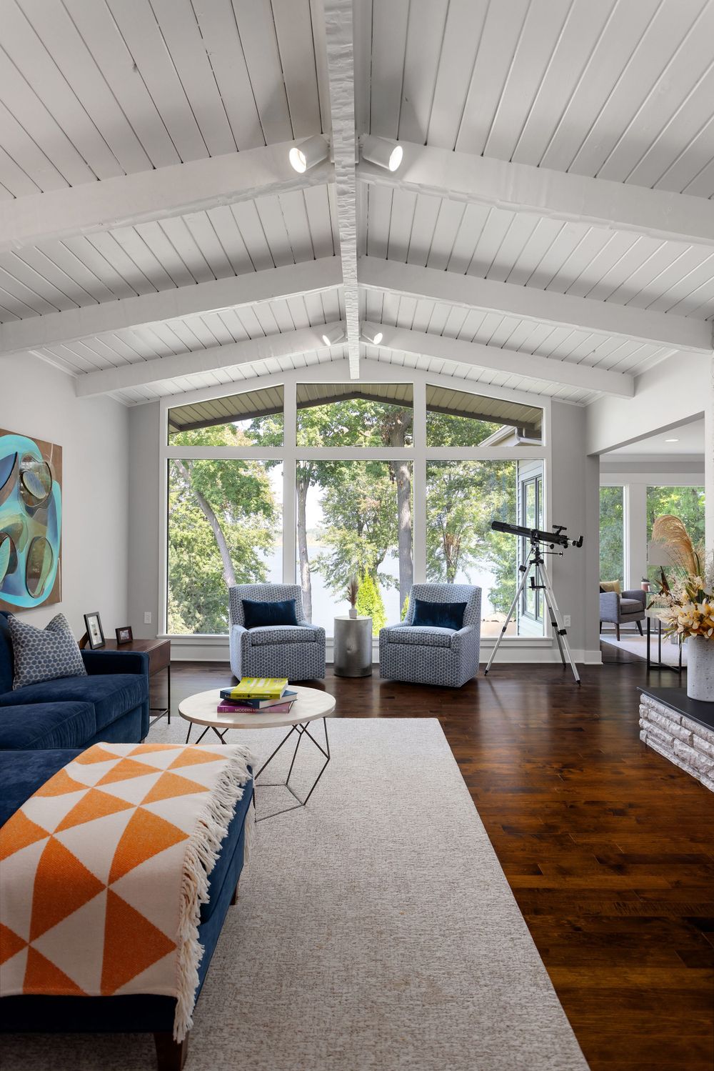 Bright living room with vaulted white ceiling, large windows overlooking a lake, modern furnishings, and a telescope facing the water view.