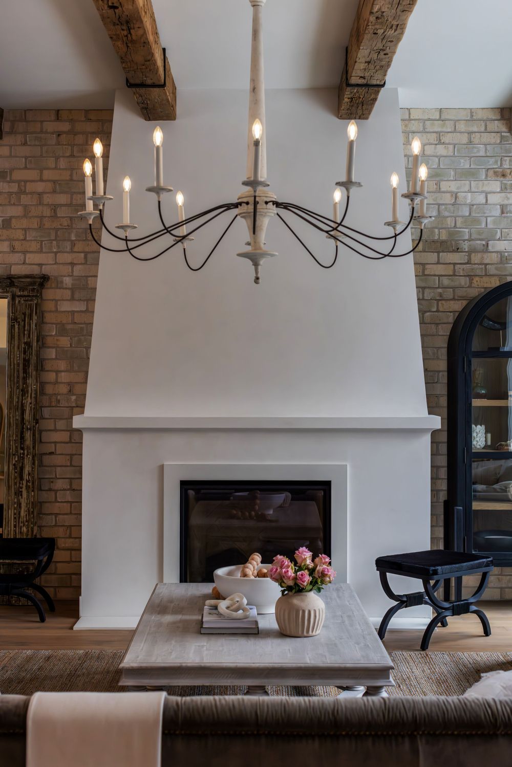 Elegant living room with rustic beams, a white plaster fireplace, modern chandelier, and a styled coffee table.
