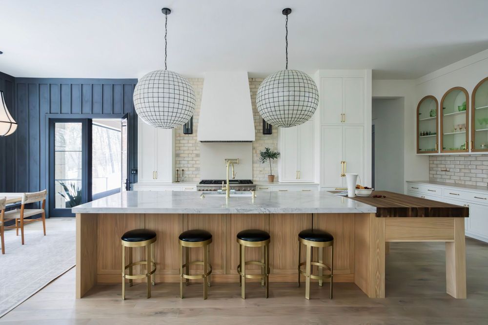 Modern luxury kitchen featuring a large marble island with a wood extension, brass & black stools, globe pendant lights