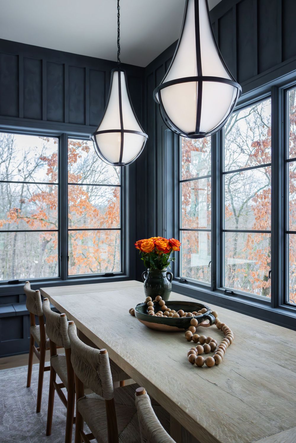 Cozy dining nook with dark paneled walls, large windows showcasing fall foliage, and a light wood table with florals and wood bead decor.