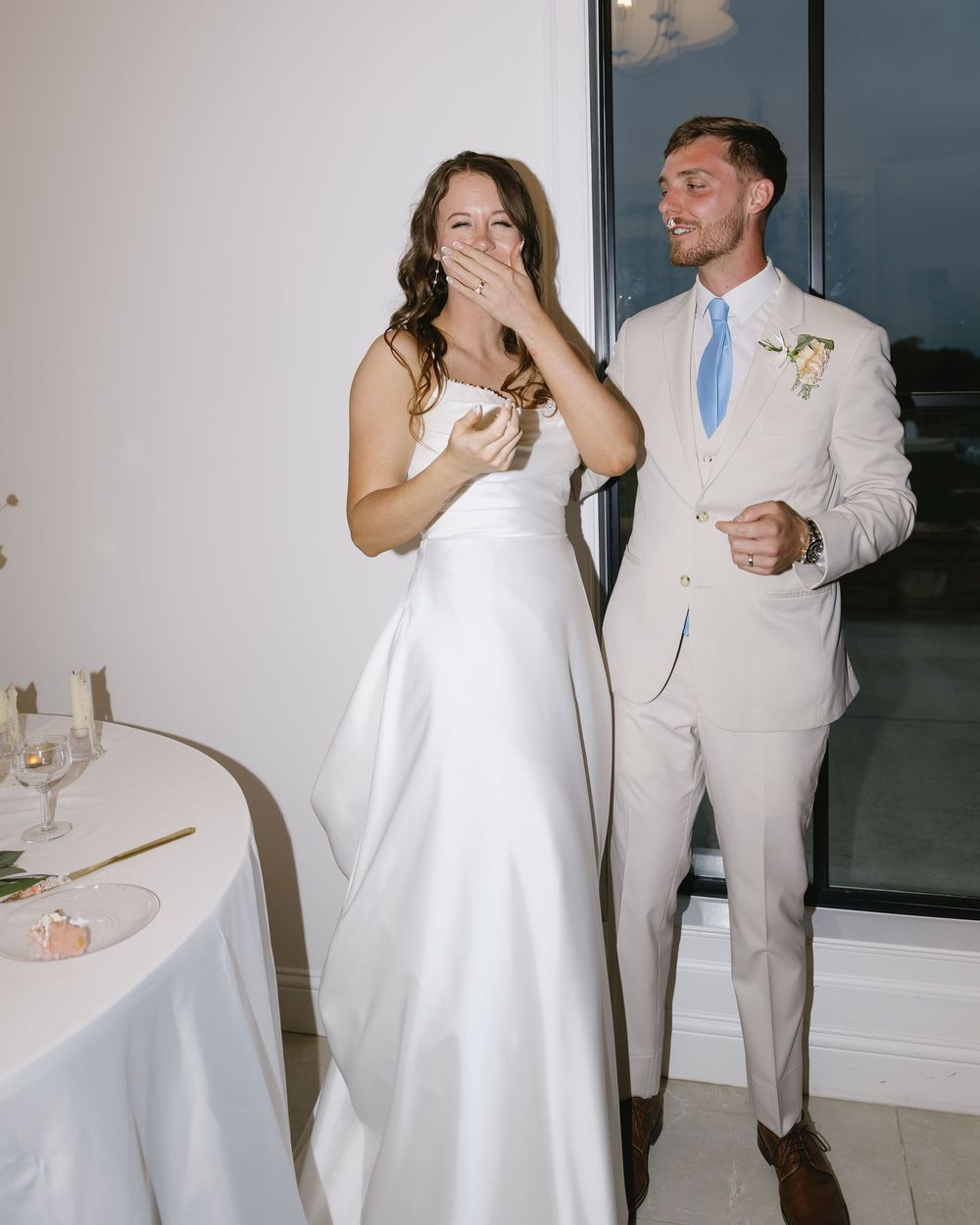 A bride in a white dress and groom in a beige suit share a candid moment during their wedding reception.