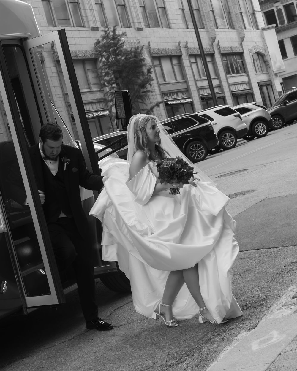 A couple exits a vehicle on their wedding day, with the bride holding her flowing white gown and bouquet.
