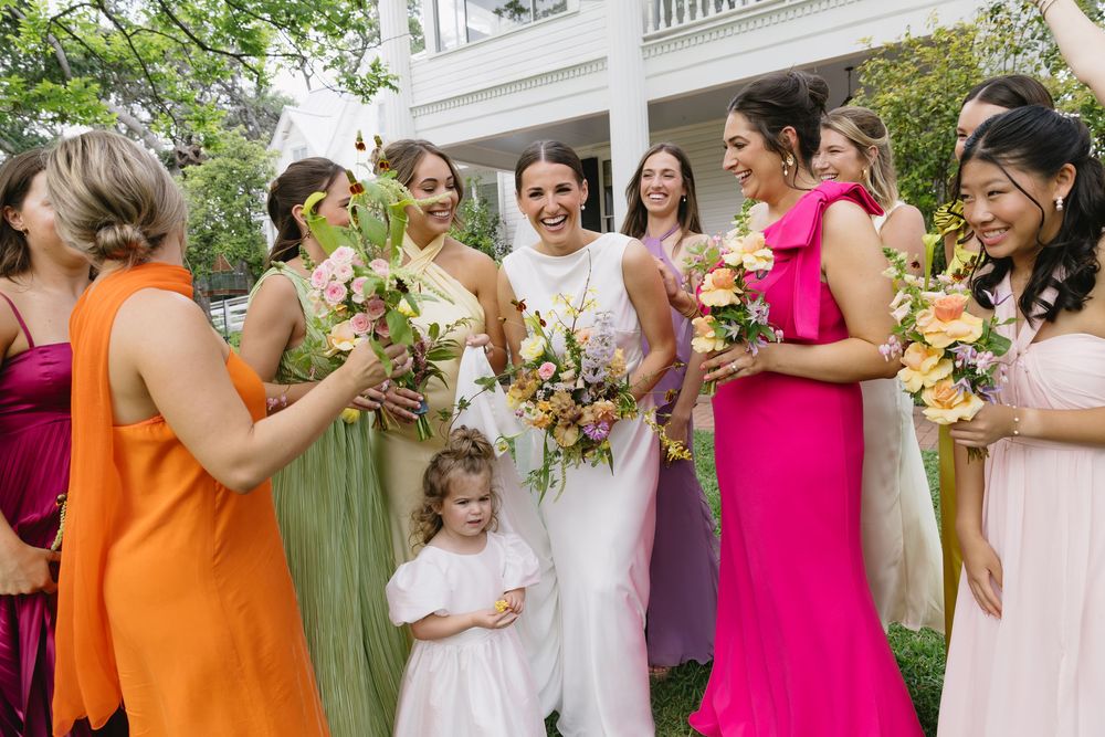 Bridal party in colorful dresses of orange, pink, and green gather outdoors with flowers for wedding photos.