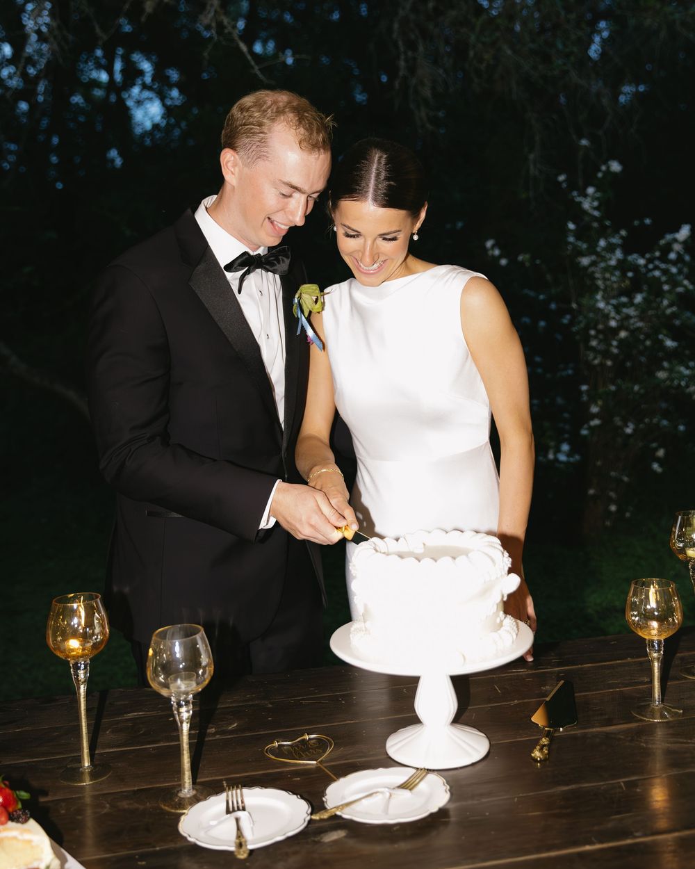 Wedding couple cutting their white tiered cake together at an evening reception with wine glasses.