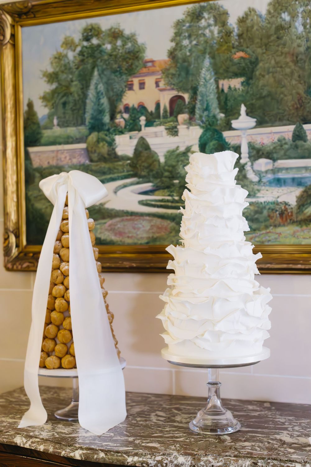 Two modern white wedding cakes displayed on marble countertop with elegant garden painting in background.