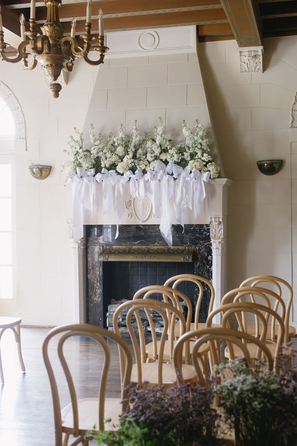Elegant wedding ceremony setup with white floral mantel arrangement above fireplace and wooden bentwood chairs.