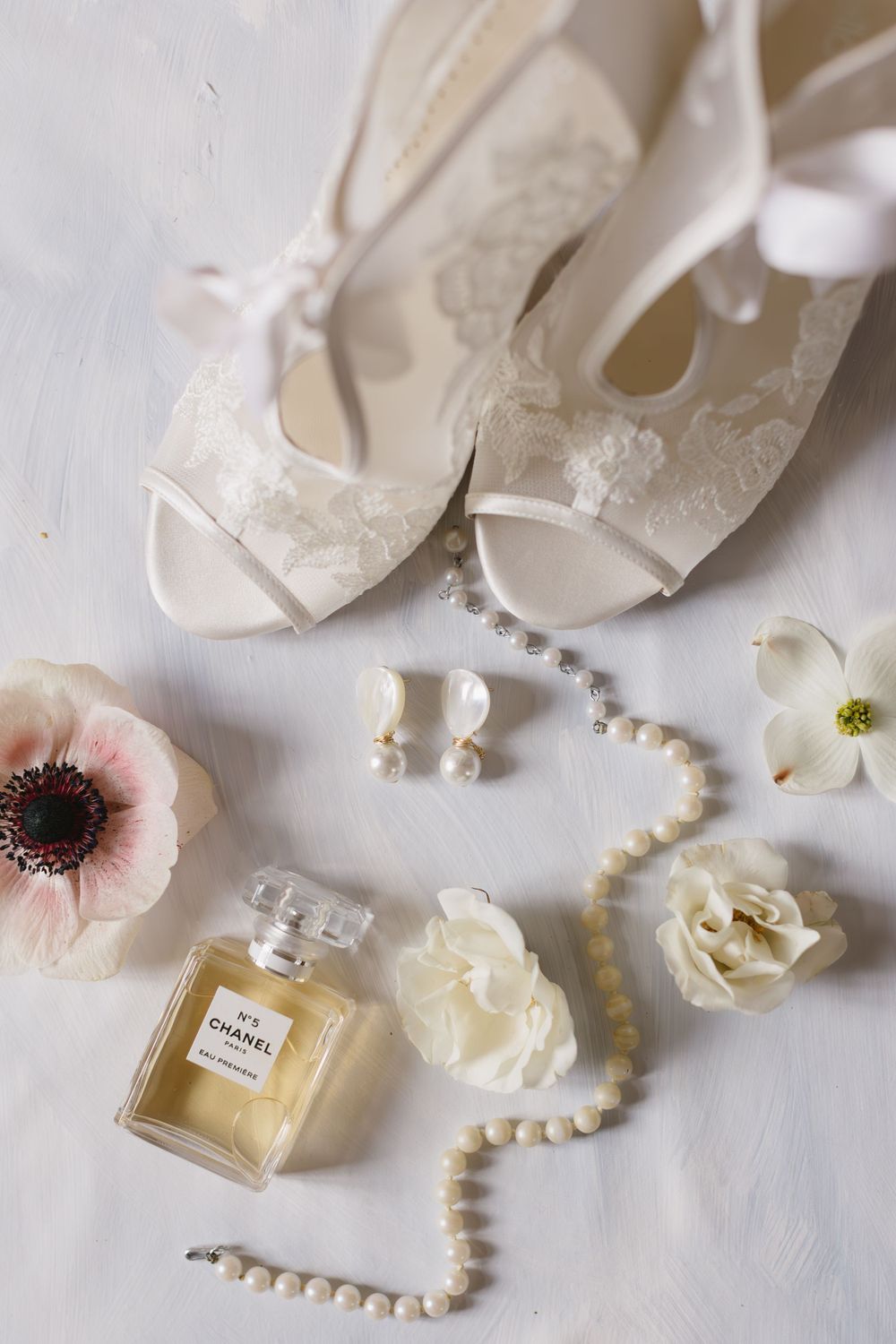 Elegant bridal accessories including white lace shoes, pearl necklace, Chanel perfume, and delicate anemone and rose blooms.