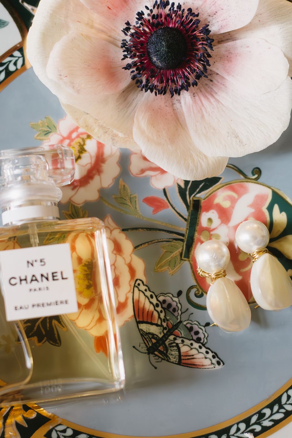 Close-up of a pink anemone flower next to a Chanel No 5 perfume bottle on an ornate plate.