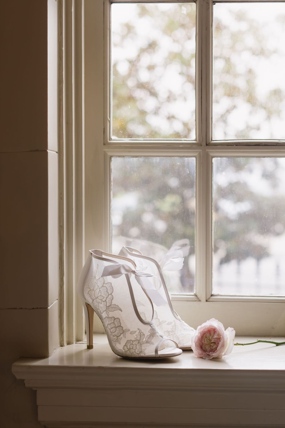 White lace bridal bootie with a pink rose on a windowsill in soft natural light.