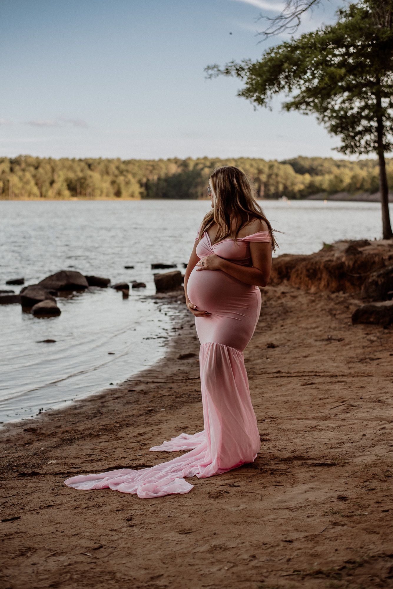 A maternity photo of someone in a flowing pink dress standing on a lakeside beach at sunset.