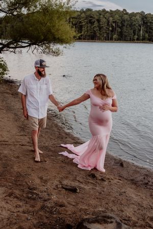 A couple walks hand in hand along a lake shore, with a flowing pink dress trailing behind.