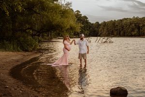 A couple stands together at the water's edge during a sunset maternity photoshoot.