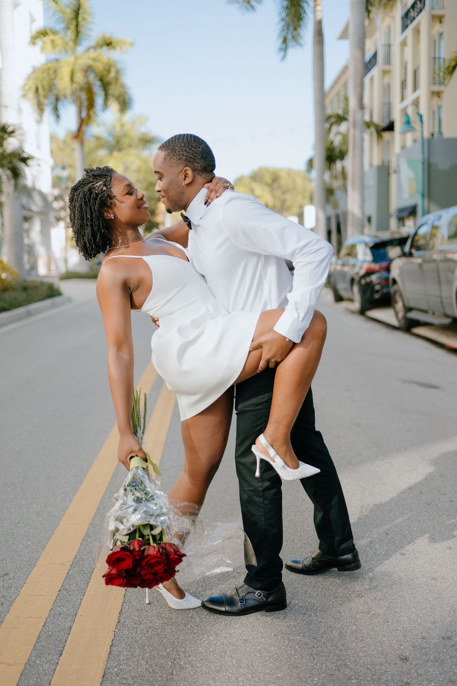 A romantic moment between a couple on a palm-lined street, with the bride holding red roses and wearing white heels.
