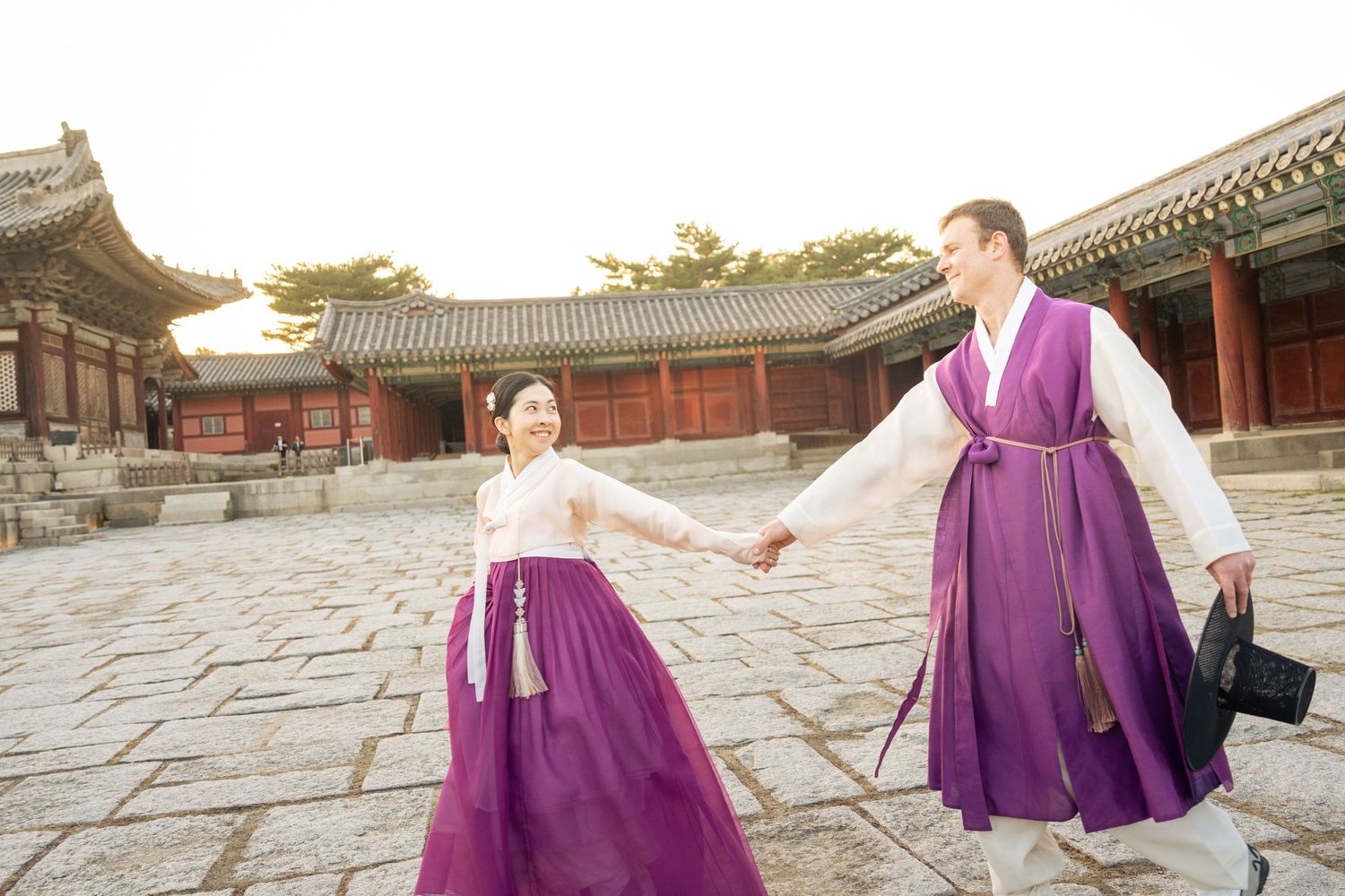 Couple in traditional purple hanbok clothing posing gracefully in Korean palace courtyard at sunset.
